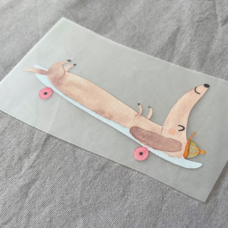 Może przedstawiać: Kresk&oacute;wkowa ilustracja przedstawiająca jamnika w kapeluszu jadącego na deskorolce. Jamnik jest brązowo-biały i ma r&oacute;żowy nos. Deskorolka jest niebieska i ma r&oacute;żowe k&oacute;łka.