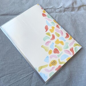 May include: A white notebook with a colorful abstract design on the cover. The design features a variety of shapes and colors, including pink, blue, green, yellow, and orange. The notebook has a clear plastic cover and the text "Happy Fine Days" is printed on the bottom right corner of the cover.