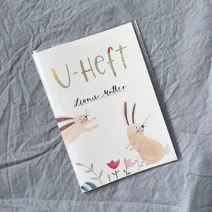 May include: A white booklet with the text "U-Heft" and "Leonie M&uuml;ller" on the front cover. The cover also features illustrations of two rabbits, one jumping and one standing, with a floral design at the bottom.