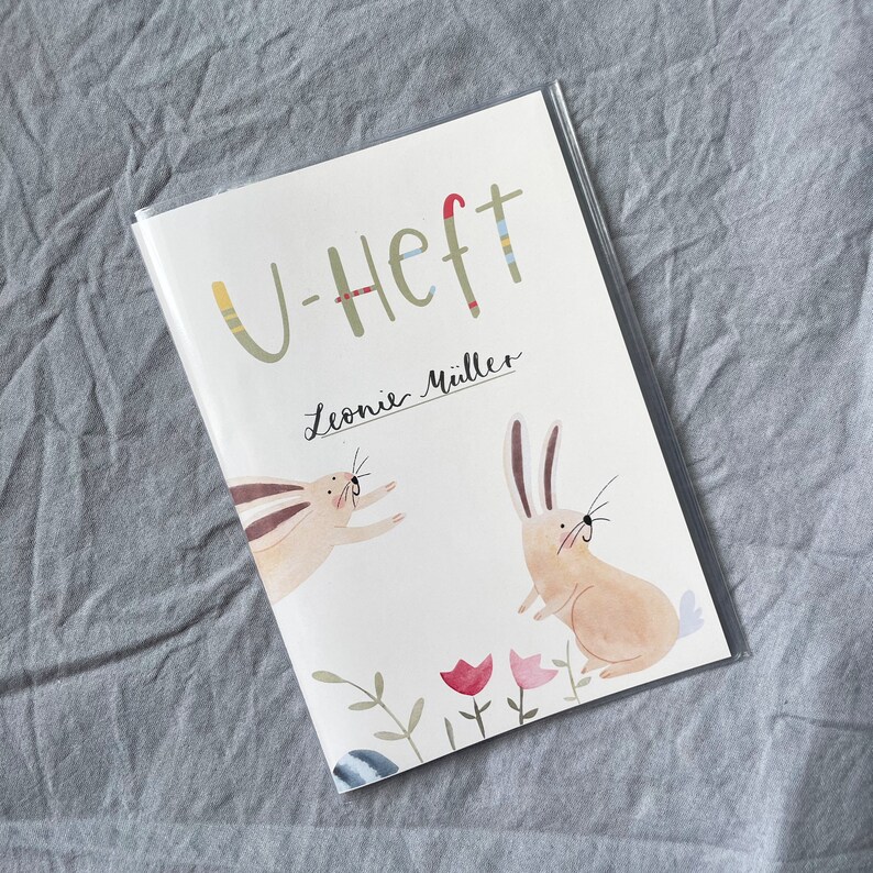 May include: A white booklet with the text "U-Heft" and "Leonie M&uuml;ller" on the front cover. The cover also features illustrations of two rabbits, one jumping and one standing, with a floral design at the bottom.
