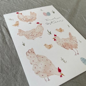 Pu&ograve; includere: Un'illustrazione ad acquerello di un biglietto con uno sfondo rosa chiaro e il testo "Frohe Ostern" in scrittura corsiva marrone. Il biglietto presenta cinque galline e pulcini, tutti con creste rosse e corpi rosa chiaro. Le galline hanno occhi marroni e macchie marrone chiaro. I pulcini hanno corpi gialli e occhi marroni. Il biglietto &egrave; decorato con erba verde e uova blu chiaro.