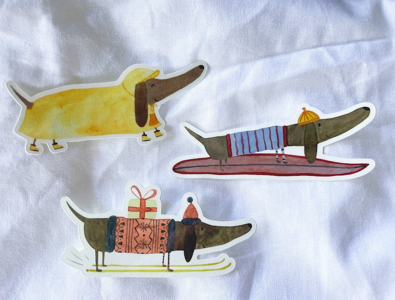 May include: Three dachshund dog stickers with different winter activities. One dachshund is wearing a yellow raincoat, another is wearing a blue and white striped shirt and is surfing, and the third is wearing a red and white sweater and is skiing with a gift.