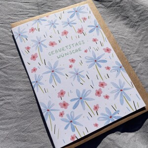 May include: A greeting card with a floral design featuring blue and pink flowers on a white background. The card has the German text "GEBURTSTAGS W&Uuml;NSCHE" and is paired with a brown envelope. The card is likely for a birthday.