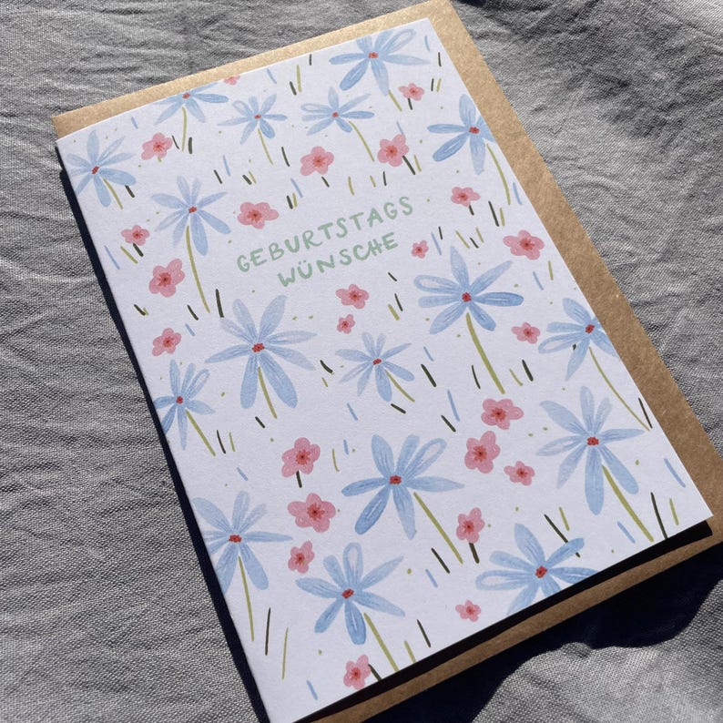 May include: A greeting card with a floral design featuring blue and pink flowers on a white background. The card has the German text "GEBURTSTAGS W&Uuml;NSCHE" and is paired with a brown envelope. The card is likely for a birthday.
