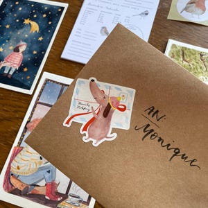 Op de afbeelding: Een bruine envelop geadresseerd aan "Monique" met een sticker van een cartoonhond met een brief. Andere items zijn een aquarel illustratie van een meisje onder een sterrenhemel, een vogel illustratie en een notitieblok met vogel illustraties.