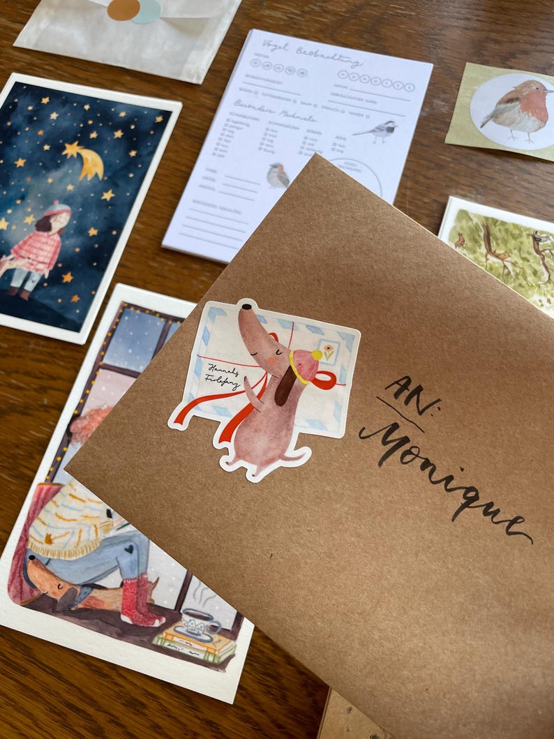 Op de afbeelding: Een bruine envelop geadresseerd aan "Monique" met een sticker van een cartoonhond met een brief. Andere items zijn een aquarel illustratie van een meisje onder een sterrenhemel, een vogel illustratie en een notitieblok met vogel illustraties.