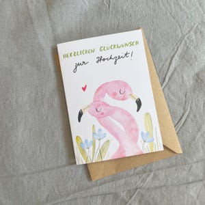May include: A wedding greeting card featuring a watercolour illustration of two pink flamingos. The card has the text "HERZLICHEN GL&Uuml;CKWUNSCH zur Hochzeit!" in green and black. It rests on a brown envelope, against a grey backdrop.