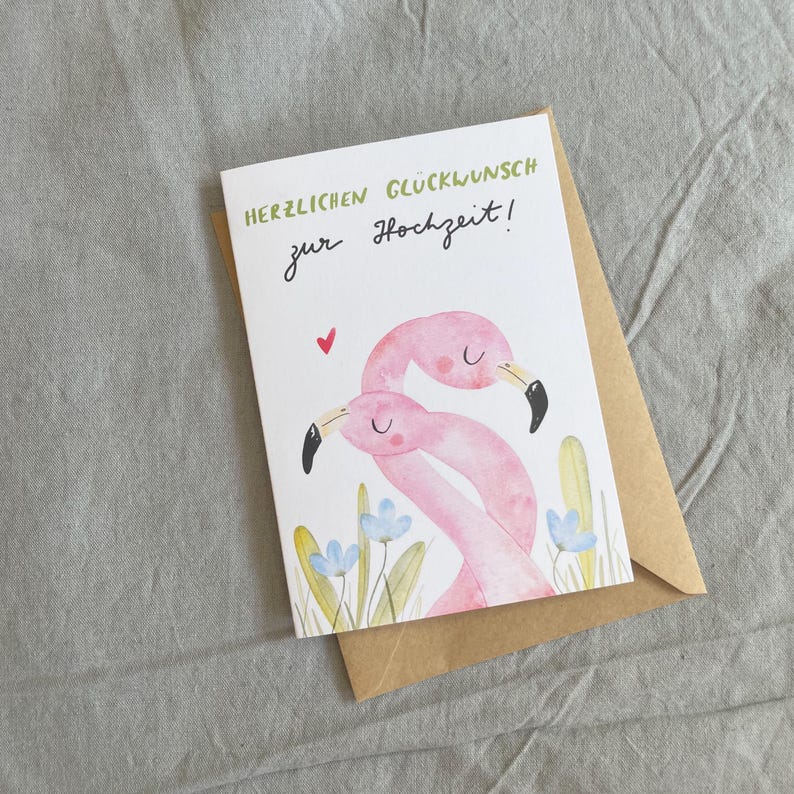 May include: A wedding greeting card featuring a watercolour illustration of two pink flamingos. The card has the text "HERZLICHEN GL&Uuml;CKWUNSCH zur Hochzeit!" in green and black. It rests on a brown envelope, against a grey backdrop.