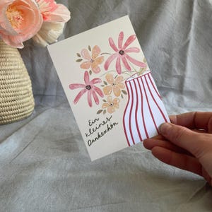 May include: A watercolour greeting card with a floral design. The card shows pink and peach flowers in a vase with red and white stripes. The card has the German text "Ein kleines Dankesch&ouml;n".