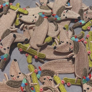 May include: Embroidered patches featuring a cartoon dog riding a skateboard. The patches are primarily tan with brown ears and accents, and the skateboards are green. The dogs have colorful hats and are scattered across a gray surface.