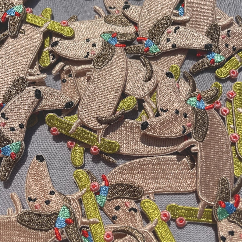 May include: Embroidered patches featuring a cartoon dog riding a skateboard. The patches are primarily tan with brown ears and accents, and the skateboards are green. The dogs have colorful hats and are scattered across a gray surface.