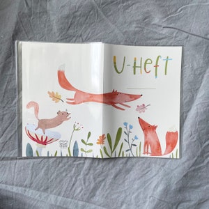 Może przedstawiać: Otwarta książka z fantazyjną ilustracją lis&oacute;w i wiewi&oacute;rki w leśnym otoczeniu. Okładka zawiera słowo "U-Heft" w kolorowych literach. Grafika zawiera rośliny, kwiaty i grzyb.