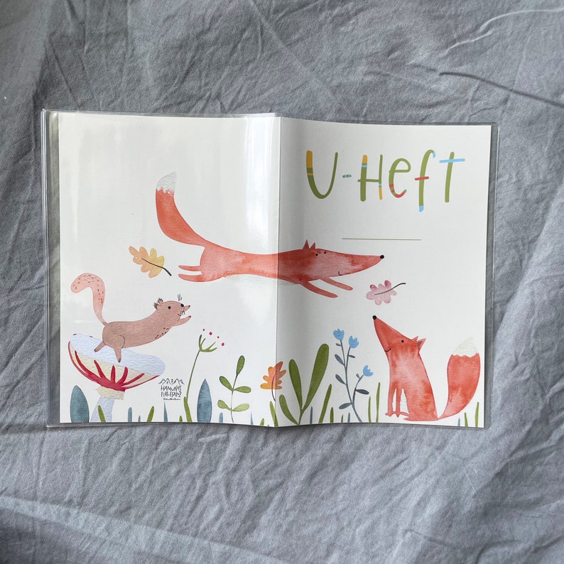 Może przedstawiać: Otwarta książka z fantazyjną ilustracją lis&oacute;w i wiewi&oacute;rki w leśnym otoczeniu. Okładka zawiera słowo "U-Heft" w kolorowych literach. Grafika zawiera rośliny, kwiaty i grzyb.