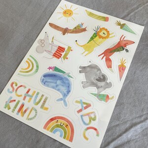 Pu&ograve; includere: Un foglio di adesivi colorati con illustrazioni stravaganti di animali, tra cui un bassotto, un leone, una volpe e un elefante. Gli adesivi includono anche un sole, una matita, un arcobaleno e le parole "SCHUL KIND" e "ABC".