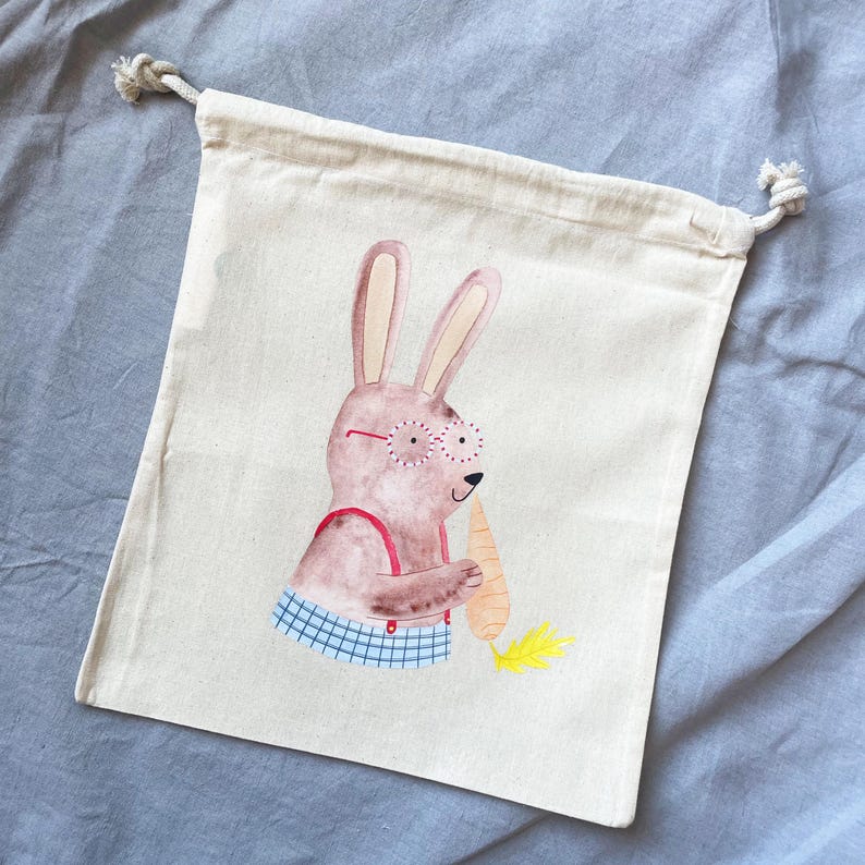May include: A white cotton drawstring bag with a cartoon illustration of a brown bunny wearing glasses and holding a carrot. The bunny is wearing a red and white checkered shirt.