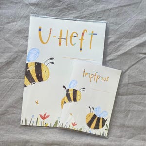 May include: Two rectangular protective covers for documents, featuring a cream background with illustrations of yellow and black striped bees, blue wings, and small colorful flowers. The larger cover displays the text U-Heft, and the smaller cover displays the text Impfpass in a colorful, playful font.