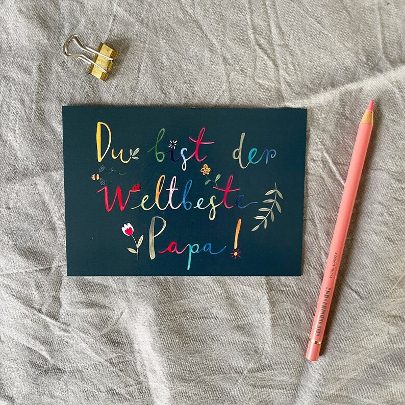 May include: A dark blue card with the text "Du bist der Weltbeste Papa!" in colorful letters. The card is decorated with small flowers, leaves, and bees.