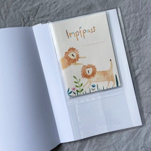 K&ouml;nnte beinhalten: Ein wei&szlig;er Reisepass-Etui mit einer durchsichtigen Plastikh&uuml;lle. Die H&uuml;lle zeigt eine bunte Illustration von zwei L&ouml;wen und das Wort "Impfpass" ist darauf gedruckt.