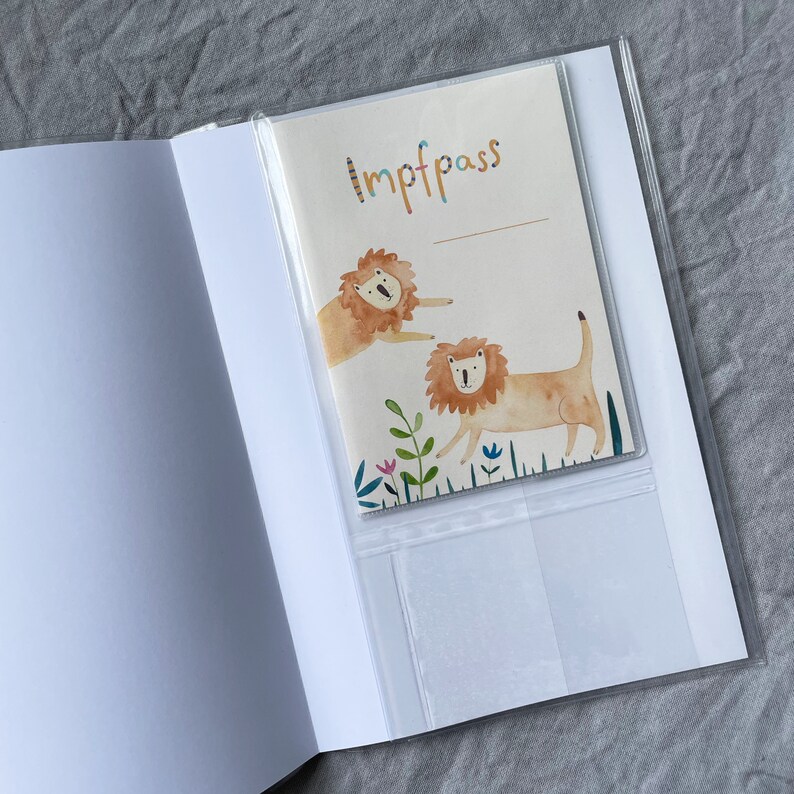 K&ouml;nnte beinhalten: Ein wei&szlig;er Reisepass-Etui mit einer durchsichtigen Plastikh&uuml;lle. Die H&uuml;lle zeigt eine bunte Illustration von zwei L&ouml;wen und das Wort "Impfpass" ist darauf gedruckt.