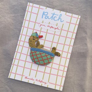 May include: An embroidered patch of a cartoon character in a blue and green basket. The character wears a blue cap with a red brim. The patch is on a card with the word "Patch" and the text "zum Aufb&uuml;geln".