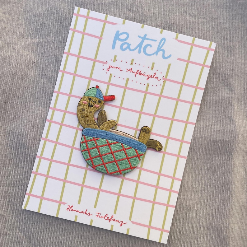 May include: An embroidered patch of a cartoon character in a blue and green basket. The character wears a blue cap with a red brim. The patch is on a card with the word "Patch" and the text "zum Aufb&uuml;geln".