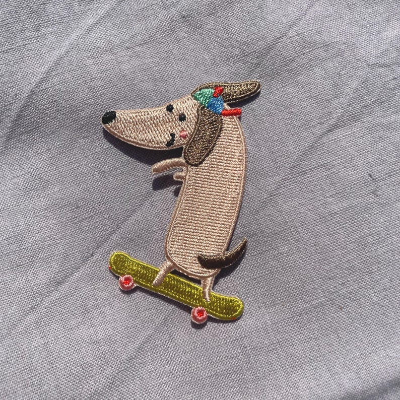 May include: Embroidered patch of a dachshund dog on a skateboard. The dog is tan with brown ears and a small hat. The skateboard is green with red wheels. The patch is designed for application on clothing or accessories.
