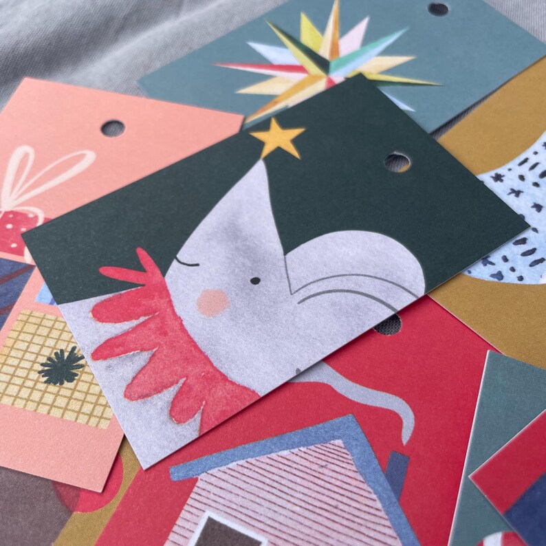 K&ouml;nnte beinhalten: Eine Sammlung farbenfroher Geschenkanh&auml;nger mit verschiedenen festlichen Designs. Die Anh&auml;nger zeigen Illustrationen eines Sterns, eines Hauses und eines skurrilen Charakters mit rotem Kragen. Jeder Anh&auml;nger hat ein kleines Loch zum Befestigen an einem Geschenk.
