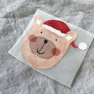 Op de afbeelding: Een bruine beer die een rode kerstmanmuts draagt met een witte pompon. De beer glimlacht en heeft een bruine neus.