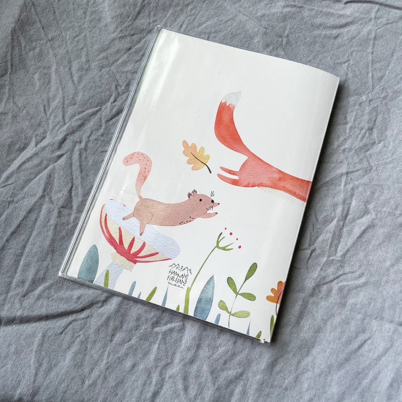 Może przedstawiać: Książka z akwarelową ilustracją lisa i wiewi&oacute;rki w leśnej scenerii. Lis jest pomarańczowy, a wiewi&oacute;rka brązowa. Książka jest w przezroczystej plastikowej okładce.