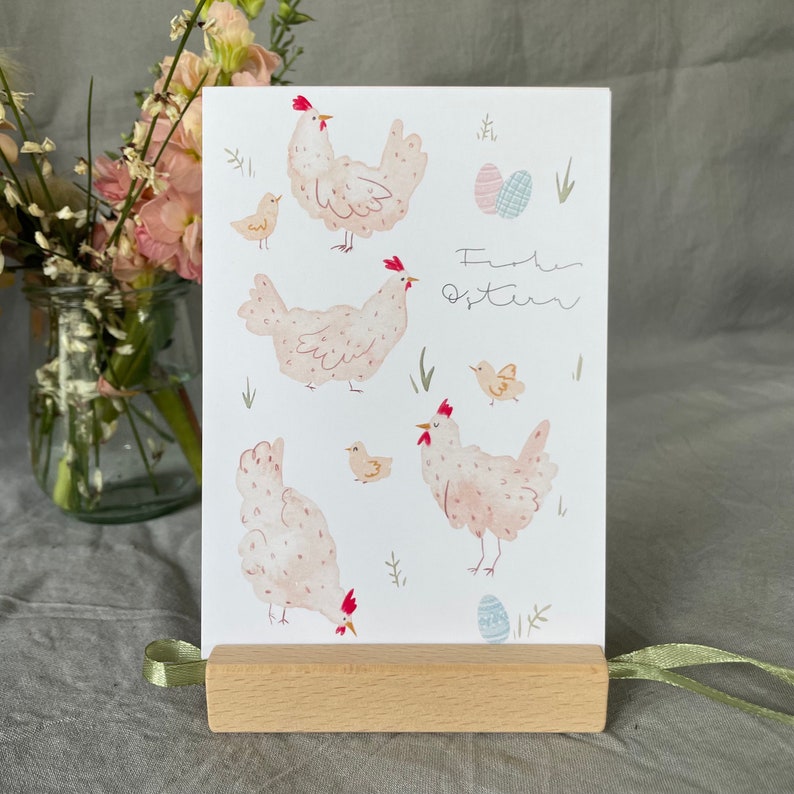 Pu&ograve; includere: Un biglietto bianco con un'illustrazione ad acquerello di galline e uova. Il biglietto dice "Frohe Ostern", che in tedesco significa "Buona Pasqua".