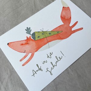 May include: A watercolour illustration of a fox wearing a green and white cone hat with the text "Auf in die Schule!" which translates to "Off to school!" in English.