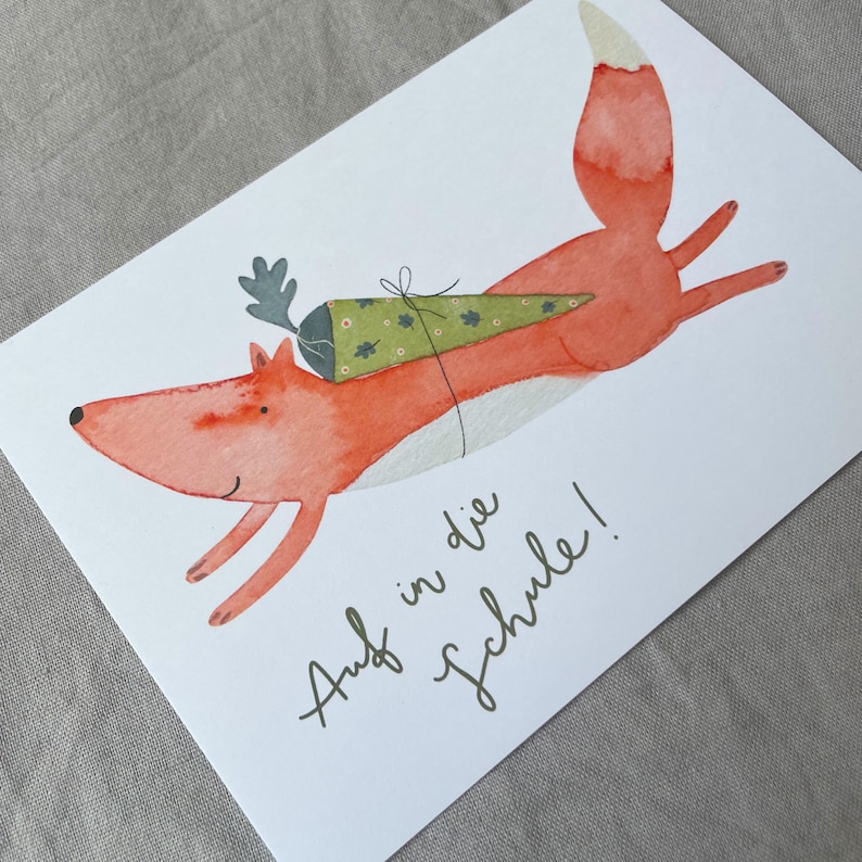 May include: A watercolour illustration of a fox wearing a green and white cone hat with the text "Auf in die Schule!" which translates to "Off to school!" in English.