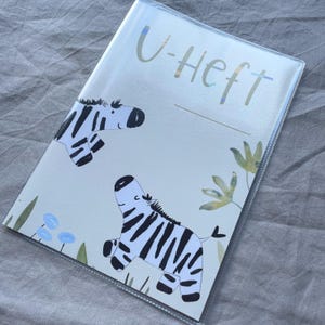 May include: A rectangular protective cover for a booklet featuring the text U-HEFT at the top. The design includes two illustrated zebras with black and white stripes, accented by small blue flowers and green foliage on a light cream background.