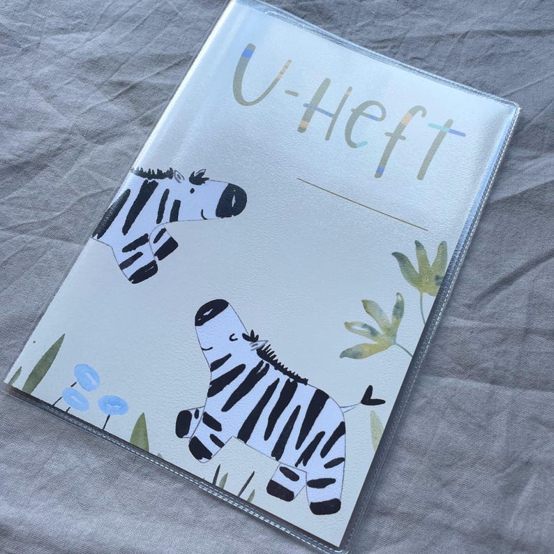 May include: A rectangular protective cover for a booklet featuring the text U-HEFT at the top. The design includes two illustrated zebras with black and white stripes, accented by small blue flowers and green foliage on a light cream background.