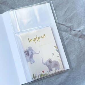 May include: A clear plastic folder with a white background and a smaller clear plastic pocket on the front. The smaller pocket contains a card with the word "Impfpass" written in cursive and a watercolour illustration of two elephants in a green and blue forest.