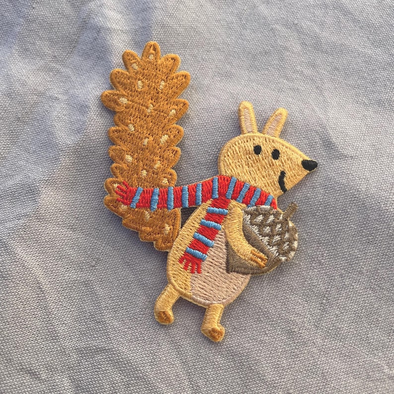 May include: Embroidered patch featuring a cartoon squirrel with a large, textured tail. The squirrel wears a red and blue striped scarf and holds a brown acorn. The patch is tan, brown, red, and blue.