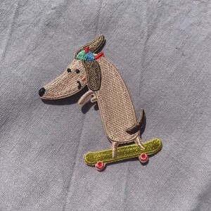 May include: Embroidered patch featuring a dachshund dog on a skateboard. The dog is light brown with a black nose, wearing a colorful hat. The skateboard is yellow with red wheels. The patch is on a gray background.