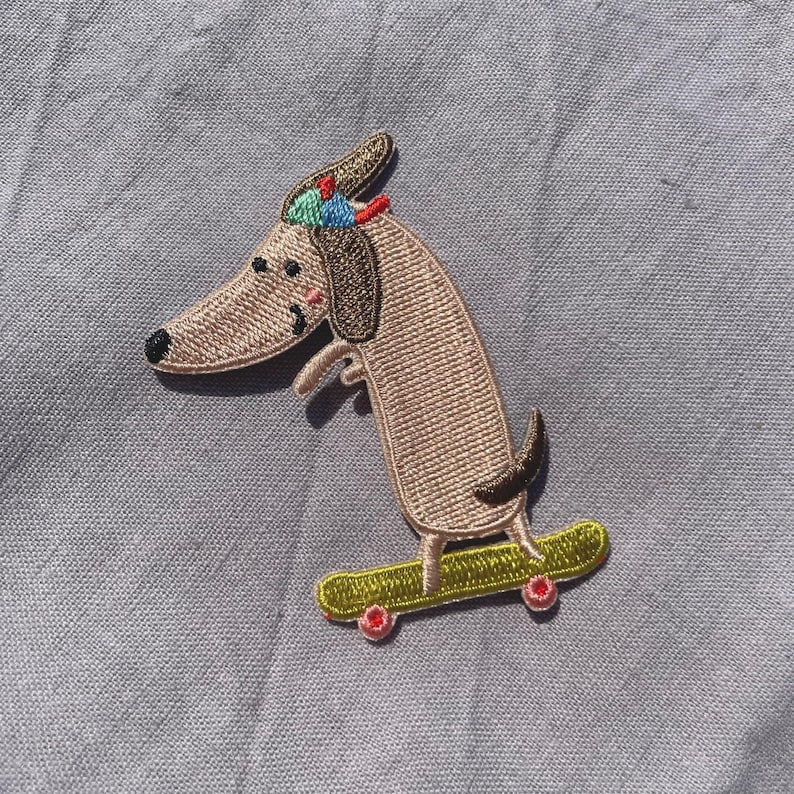 May include: Embroidered patch featuring a dachshund dog on a skateboard. The dog is light brown with a black nose, wearing a colorful hat. The skateboard is yellow with red wheels. The patch is on a gray background.
