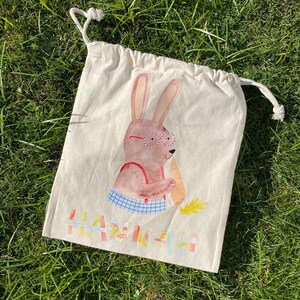 May include: A white cotton drawstring bag with a cartoon bunny wearing glasses and holding a carrot. The bag has the name "Hannah" printed on it in colorful letters.