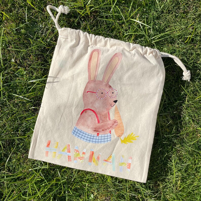May include: A white cotton drawstring bag with a cartoon bunny wearing glasses and holding a carrot. The bag has the name "Hannah" printed on it in colorful letters.