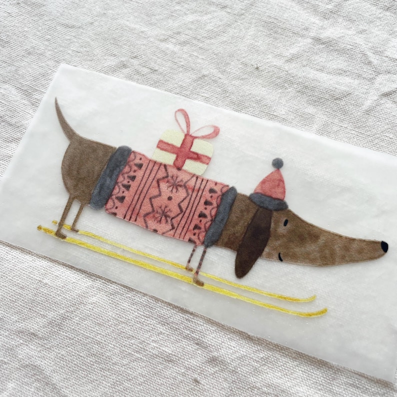 May include: A brown dachshund wearing a red and grey sweater and a red hat is carrying a wrapped gift on its back. The dachshund is on skis with yellow poles.