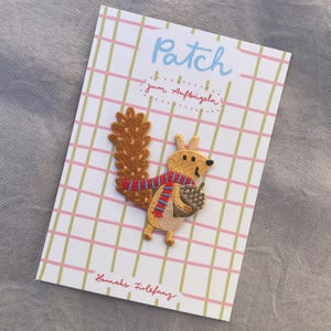 May include: Embroidered squirrel patch with a brown tail, wearing a red and blue striped scarf, and holding an acorn. The patch is on a card with the word "Patch" and "Hannahs Firlefanz" printed on it.