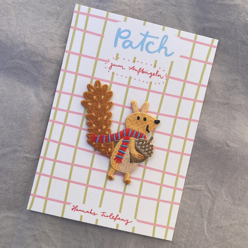 May include: Embroidered squirrel patch with a brown tail, wearing a red and blue striped scarf, and holding an acorn. The patch is on a card with the word "Patch" and "Hannahs Firlefanz" printed on it.