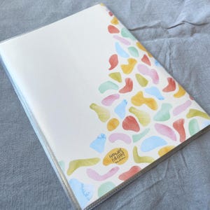 May include: A white notebook with a clear plastic cover. The cover features a colorful abstract design of overlapping shapes in shades of pink, blue, yellow, green, and orange. The text "HAMAM" and "FIRE ESCAPE" is printed in black at the bottom of the design.