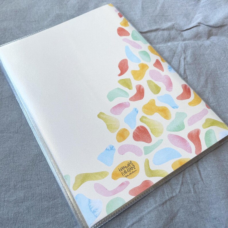 May include: A white notebook with a clear plastic cover. The cover features a colorful abstract design of overlapping shapes in shades of pink, blue, yellow, green, and orange. The text "HAMAM" and "FIRE ESCAPE" is printed in black at the bottom of the design.