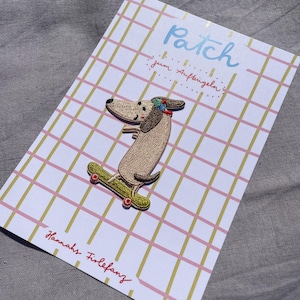 May include: Embroidered patch of a dachshund dog on a skateboard, with the word "Patch" in light blue script. The patch is on a card with a pink and gold grid pattern. The text "Hannahs Firlefanz" is written in red script.