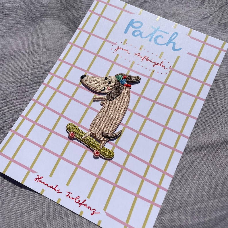 May include: Embroidered patch of a dachshund dog on a skateboard, with the word "Patch" in light blue script. The patch is on a card with a pink and gold grid pattern. The text "Hannahs Firlefanz" is written in red script.