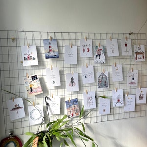 Puede incluir: Una exhibici&oacute;n de tarjetas navide&ntilde;as ilustradas, sujetas a una rejilla met&aacute;lica con pinzas de madera. Las tarjetas presentan escenas invernales, como un faro, casas y adornos festivos. Una planta colgante a&ntilde;ade un toque natural a la composici&oacute;n.