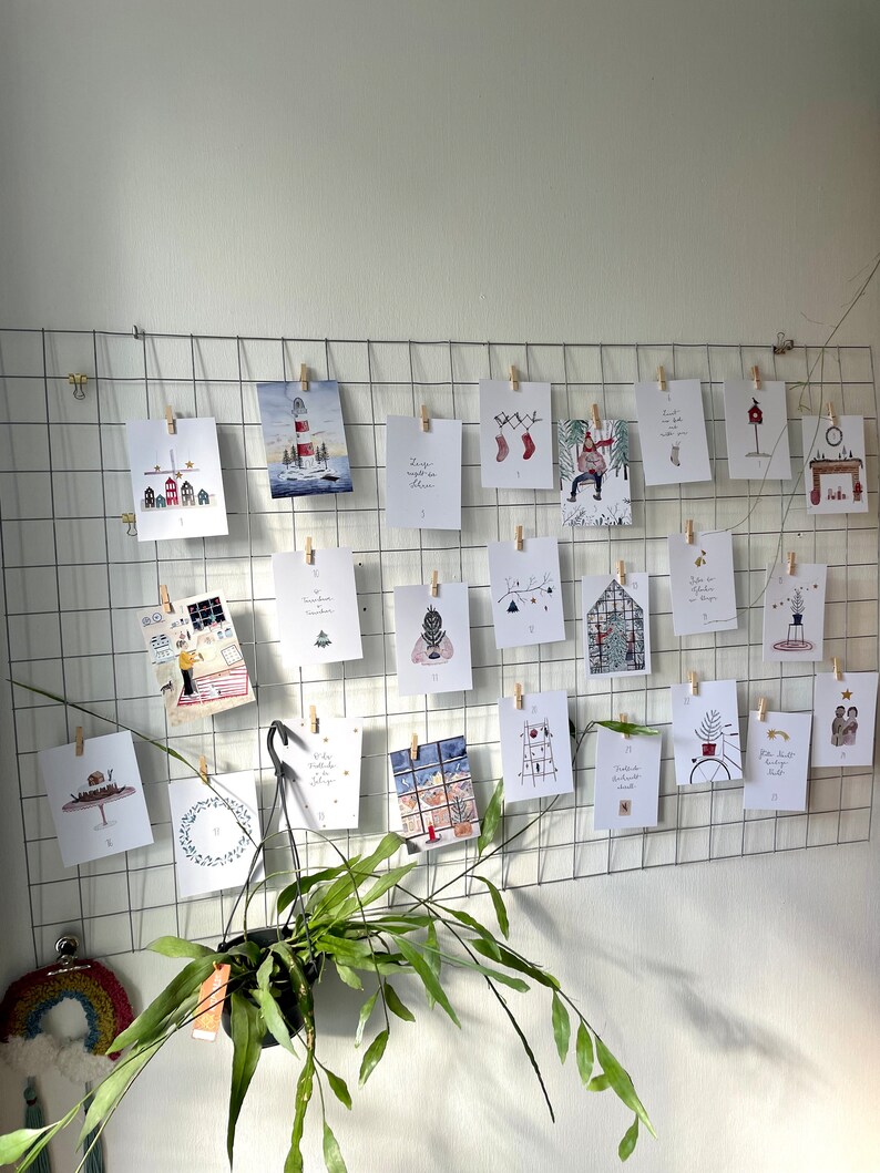 Puede incluir: Una exhibici&oacute;n de tarjetas navide&ntilde;as ilustradas, sujetas a una rejilla met&aacute;lica con pinzas de madera. Las tarjetas presentan escenas invernales, como un faro, casas y adornos festivos. Una planta colgante a&ntilde;ade un toque natural a la composici&oacute;n.