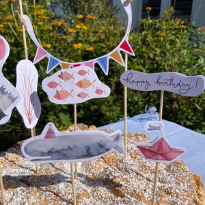 May include: A birthday cake decorated with a whale, a paper boat, and a string of colorful flags. The cake is topped with powdered sugar and the words "happy birthday!" are written on a banner.
